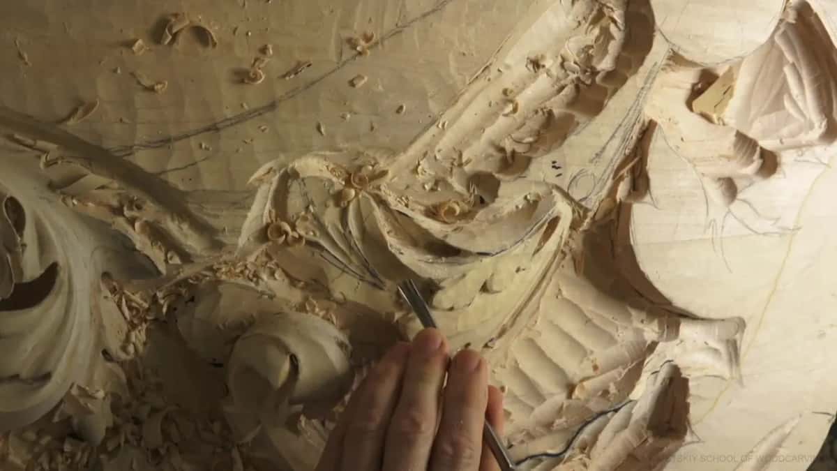 One hand pushes a gouge low in the carving, under the curled scroll