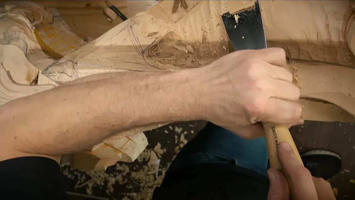 A broad flat gouge with a pale handle cleans the surface near a drawn scroll