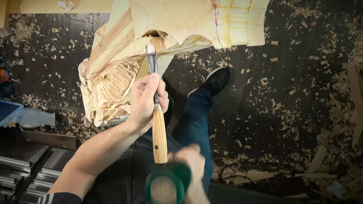 Seen from above, a mallet drives a gouge into the drop of the big carved block