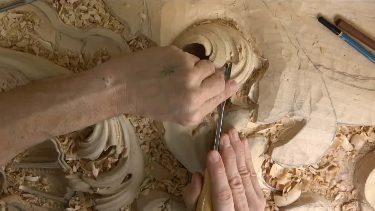 From the left, a gouge dips into the curl while two pencils rest on the flat wood above.