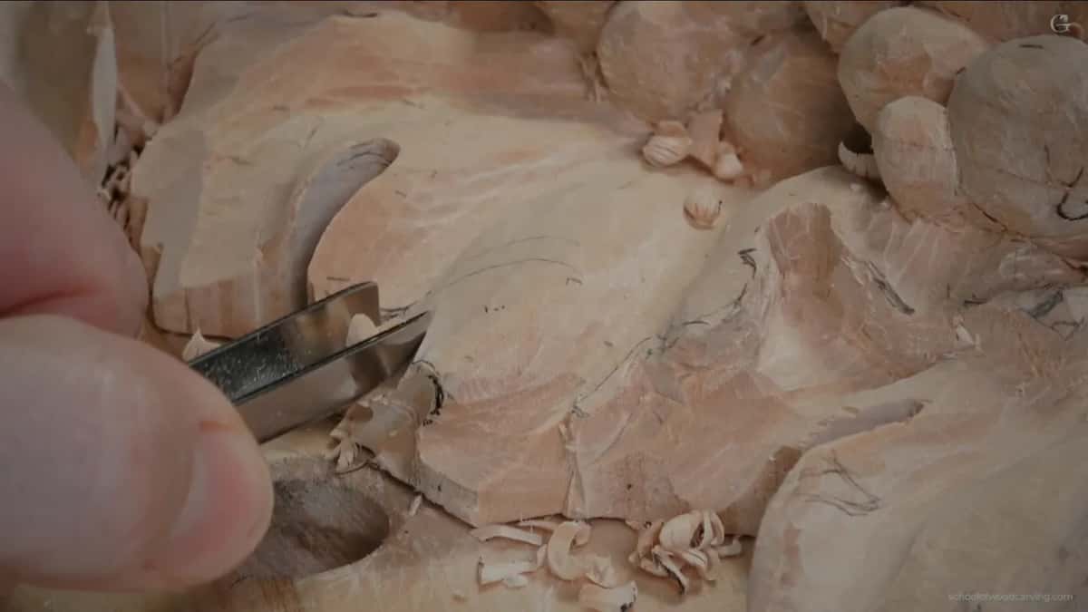 Wide gouge with a bright blade pushing into the edge of the leaf, curled chips scattered on the board
