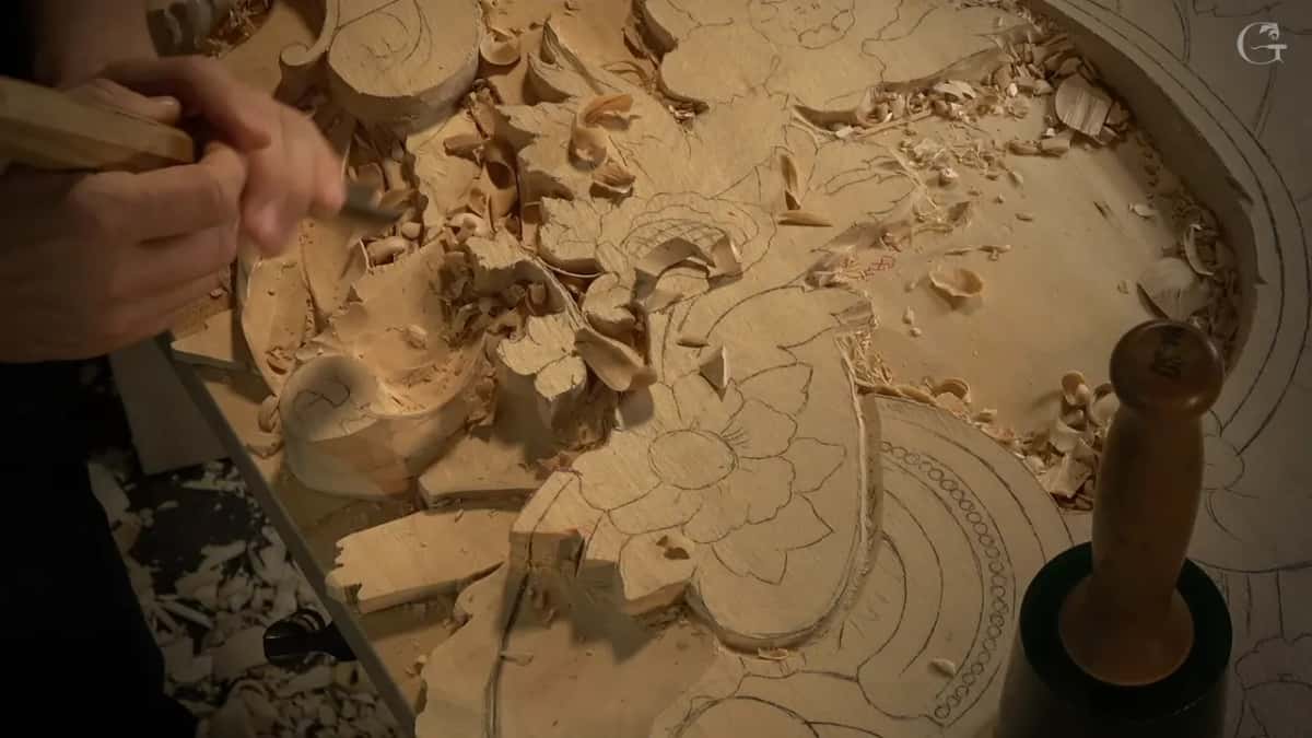 The whole corner from above, chips scattered, one hand working a long tool beside the scroll while the mallet stands by
