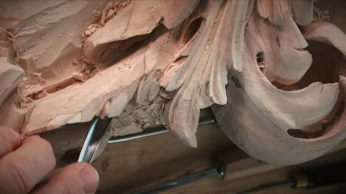 Gouge coming in low beneath a carved leaf, a big scroll to the right, tools resting on the bench below