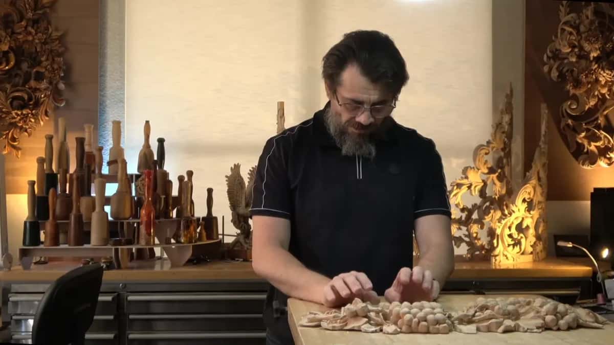 Alexander at his bench with both hands on the long grape panel, gilded carvings glowing on the wall behind him.