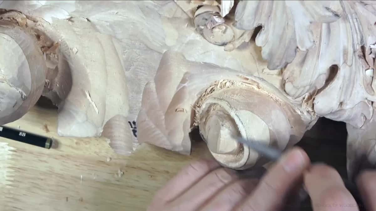 Near a pencil lying on the bench, both hands work a tool along the lower right side of the curl.