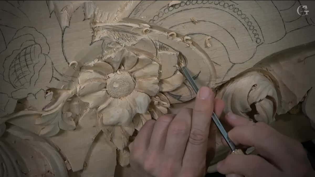 Two hands guide one narrow gouge beside a carved flower with a textured center
