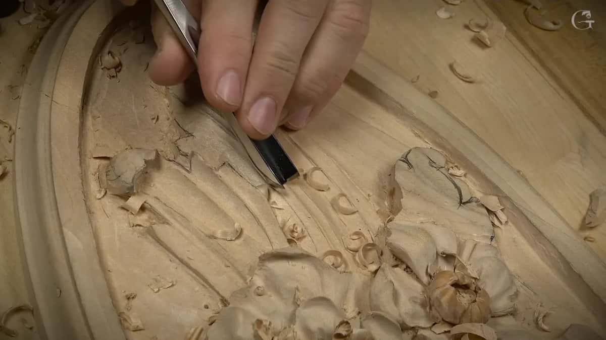 Fingers guide a gouge down a long shallow groove, the flowers off to one side
